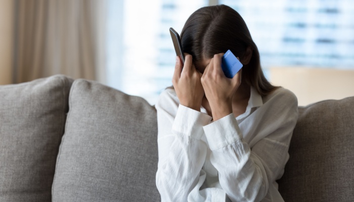 A woman holds her head with a phone in one hand and a bank card in the other.